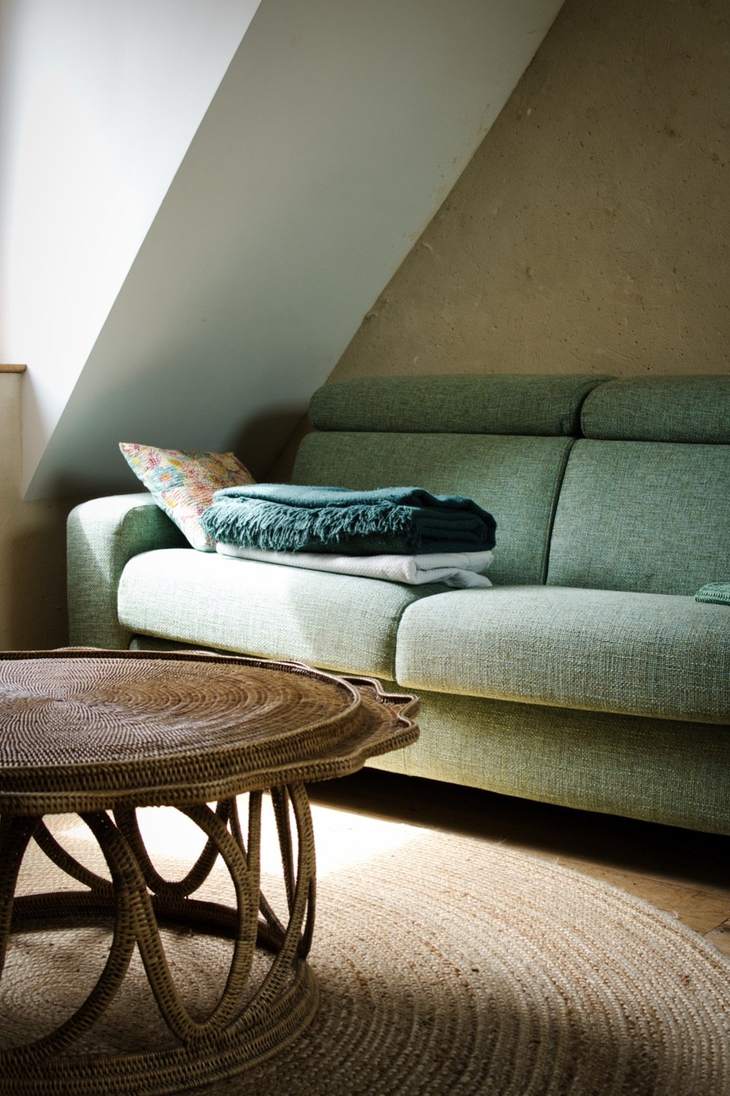 Canapé vert et table basse en rotin sous un plafond mansardé